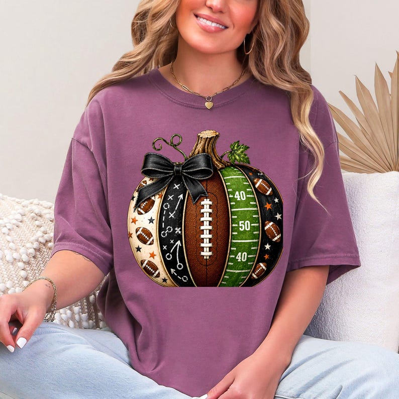 May include: A mauve short-sleeve t-shirt featuring a graphic of a pumpkin designed with football elements. The pumpkin includes a black bow, a brown football texture, a green field pattern with yard lines, and small football and star icons.