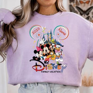 May include: A light purple sweatshirt featuring a colorful Disney graphic. The graphic includes Mickey and Minnie Mouse, Goofy, Donald Duck, and other characters. The word "Disney" is displayed with "Family Vacation" below. The year "2026" is also visible.