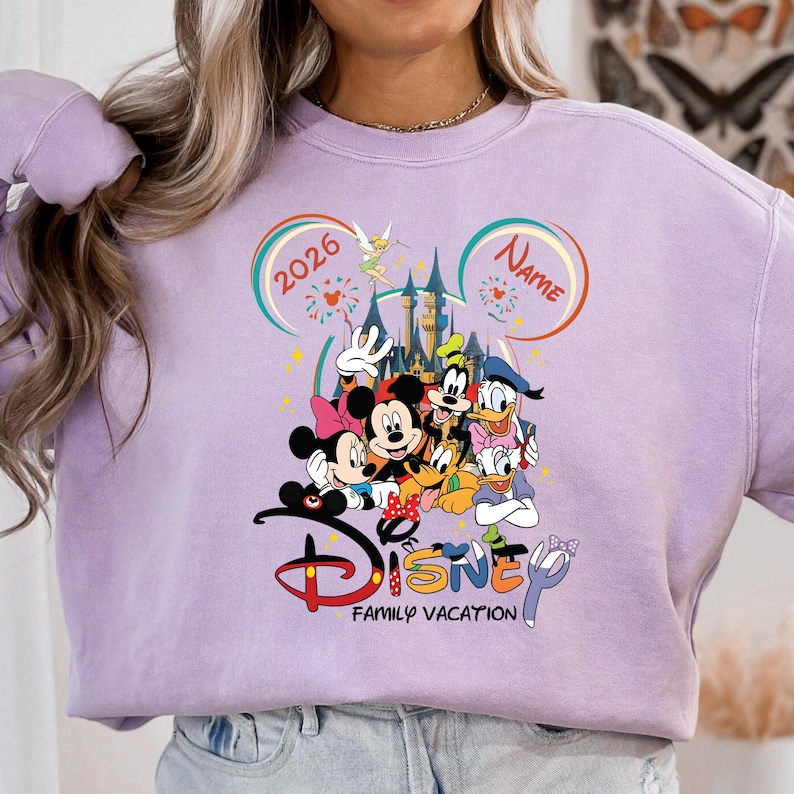 May include: A light purple sweatshirt featuring a colorful Disney graphic. The graphic includes Mickey and Minnie Mouse, Goofy, Donald Duck, and other characters. The word "Disney" is displayed with "Family Vacation" below. The year "2026" is also visible.