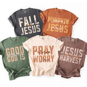 May include: Five short-sleeve crewneck t-shirts in charcoal, burnt orange, olive green, cream, and brown. Each shirt features distressed-style block lettering with religious phrases: FALL FOR JESUS, PUMPKIN SPICE JESUS, GOOD GOD IS ALL THE TIME, PRAY MORE WORRY LESS, and JESUS IS MY HARVEST.