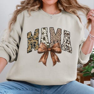 May include: A beige sweatshirt with the word "MAMA" in a collegiate font. The letters have a camouflage, leopard print, and cowhide pattern. A large brown bow is centered below the word. The sweatshirt is a casual style.