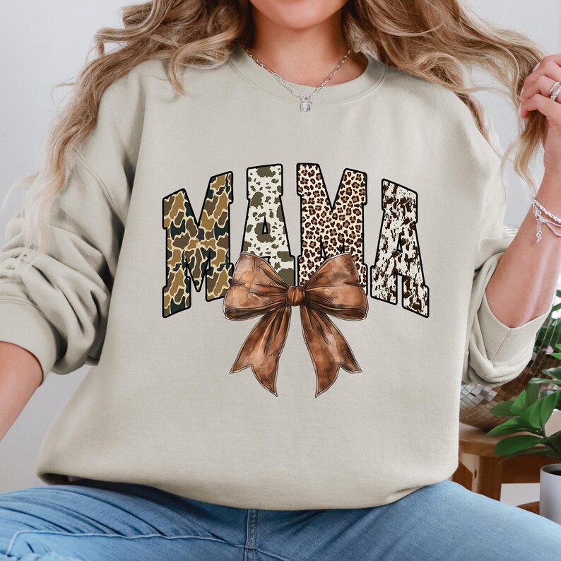 May include: A beige sweatshirt with the word "MAMA" in a collegiate font. The letters have a camouflage, leopard print, and cowhide pattern. A large brown bow is centered below the word. The sweatshirt is a casual style.