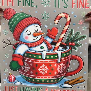Op de afbeelding: Een illustratie toont een sneeuwpop met een rode en groene muts, sjaal en wanten in een rode mok. De mok heeft sneeuwvlokontwerpen en de tekst "I'M FINE, IT'S FINE, JUST HAVING A MELTDOWN". Een zuurstok en kaneelstokjes zijn ook aanwezig.