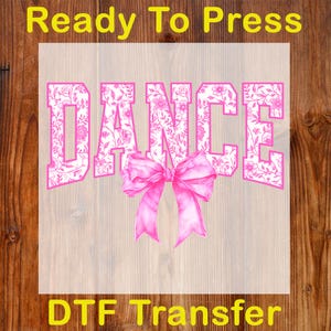 May include: A pink DTF transfer with the word "DANCE" in a bold, floral-patterned font. A pink bow is centered below the word. The text "Ready To Press" and "DTF Transfer" are in yellow.