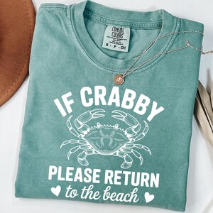 Puede incluir: Camiseta verde mar con el texto "IF CRABBY PLEASE RETURN to the beach" y un dibujo de cangrejo blanco. Un collar dorado con un colgante est&aacute; sobre la camiseta. La camiseta est&aacute; doblada y expuesta sobre una superficie blanca.