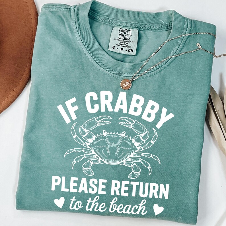 Puede incluir: Camiseta verde mar con el texto "IF CRABBY PLEASE RETURN to the beach" y un dibujo de cangrejo blanco. Un collar dorado con un colgante est&aacute; sobre la camiseta. La camiseta est&aacute; doblada y expuesta sobre una superficie blanca.