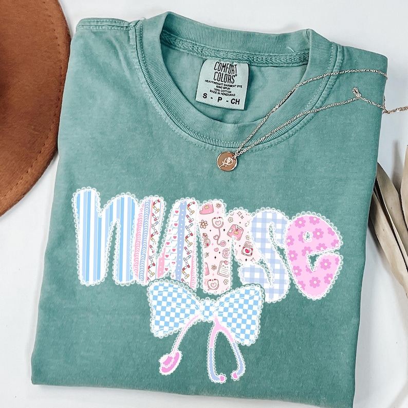 May include: A seafoam green Comfort Colors t-shirt with the word "NURSE" in a decorative font. The letters are in various patterns, including blue stripes, pink patterns, and gingham. A silver necklace with a pendant is also visible.