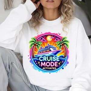 May include: White sweatshirt featuring a colorful graphic of a cruise ship on the ocean with palm trees and the words "CRUISE MODE ACTIVATED". The design is in a vibrant, neon style, perfect for vacation.