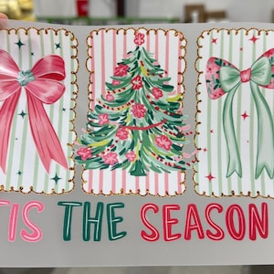 Op de afbeelding: Een kerstontwerp met drie rechthoekige frames met een gouden glitterrand. De frames tonen een roze strik, een kerstboom en een groene strik. De tekst "TIS THE SEASON" is geschreven in roze en groen.