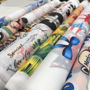 May include: A collection of rolled fabric featuring various designs. Some rolls display cartoon characters, floral patterns, and patriotic themes with red, white, and blue colors. Other rolls have text that reads "Special, Lovely, Unique" and "God".