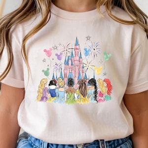 以下が含まれることがあります： ピンク色のTシャツ。水彩画で描かれたピンク色の城、花火、ディズニーのプリンセスキャラクターのイラスト。カラフルな風船と星もデザインに含まれています。