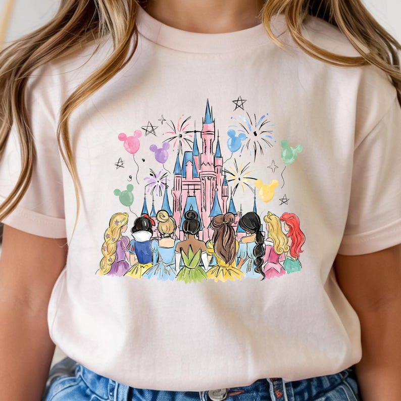 以下が含まれることがあります： ピンク色のTシャツ。水彩画で描かれたピンク色の城、花火、ディズニーのプリンセスキャラクターのイラスト。カラフルな風船と星もデザインに含まれています。