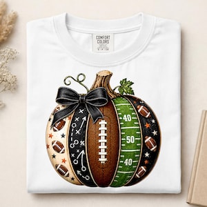 May include: A white t-shirt featuring a graphic of a pumpkin designed with American football elements. The pumpkin includes a brown football texture, a green field section with yard lines, a black play diagram, and a black bow on top.