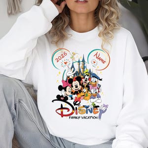 May include: White sweatshirt featuring a colorful Disney family vacation design. The graphic includes Mickey and Minnie Mouse, Goofy, Donald Duck, and other characters. The design also includes the text "Disney Family Vacation" and the year "2026".