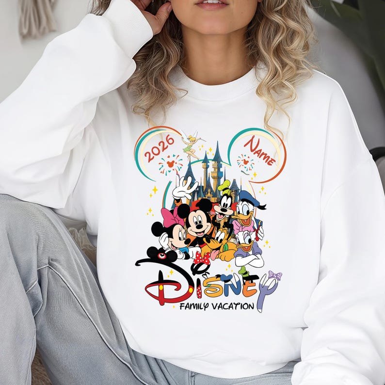 May include: White sweatshirt featuring a colorful Disney family vacation design. The graphic includes Mickey and Minnie Mouse, Goofy, Donald Duck, and other characters. The design also includes the text "Disney Family Vacation" and the year "2026".