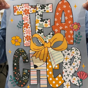 Op de afbeelding: Een transparante folie met het woord "TEACHER" in grote, kleurrijke letters. De letters zijn versierd met bloempatronen, stippen en strepen. In het midden bevinden zich een gele strik en een potlood.