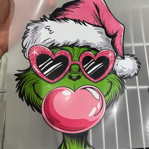 Op de afbeelding: Een groen stripfiguur met een roze kerstmuts en hartvormige zonnebril met een roze kauwgomblaas. Het personage heeft een witte bontrand op de hoed en een roze kauwgomblaas. De zonnebril heeft een glinsterend roze montuur.