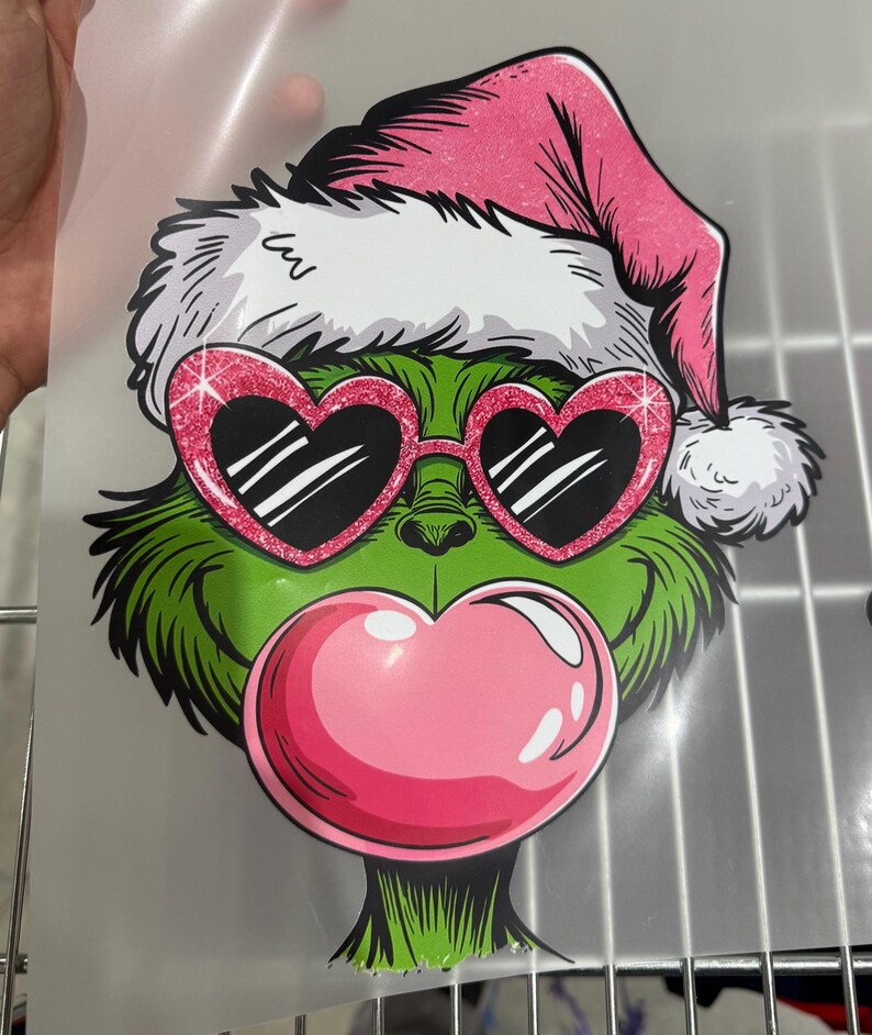Op de afbeelding: Een groen stripfiguur met een roze kerstmuts en hartvormige zonnebril met een roze kauwgomblaas. Het personage heeft een witte bontrand op de hoed en een roze kauwgomblaas. De zonnebril heeft een glinsterend roze montuur.