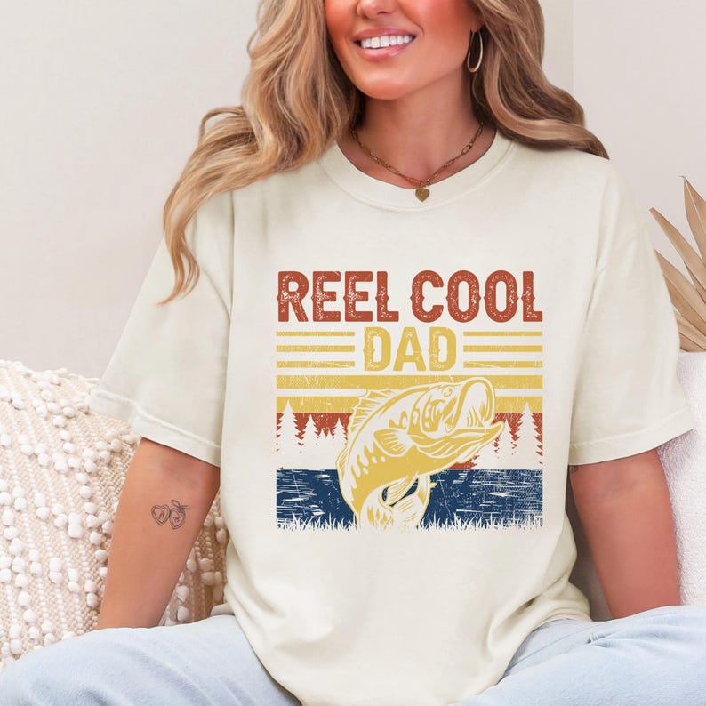 May include: Cream-colored t-shirt with the text "REEL COOL DAD" in a retro font. A graphic of a fish jumping out of the water is below the text. The design incorporates shades of orange, yellow, and blue, evoking a vintage fishing theme.