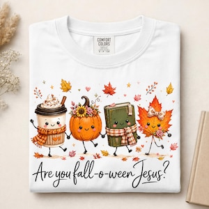 May include: A white t-shirt featuring a graphic of a coffee cup, a pumpkin, a book, and a maple leaf, all with smiling faces and limbs. The text below reads, "Are you fall-o-ween Jesus?" The shirt has a Comfort Colors brand label at the neck.