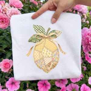 May include: White sweatshirt featuring a patchwork lemon design. The lemon is composed of various fabrics in yellow, green, and floral patterns, with a bow and leaves at the top. The design is centered on the front of the sweatshirt.