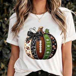 May include: A white t-shirt featuring a graphic of a pumpkin designed with football-themed elements. The pumpkin includes sections with football laces, a green field with yard lines, play diagrams, and small footballs, topped with a black bow.