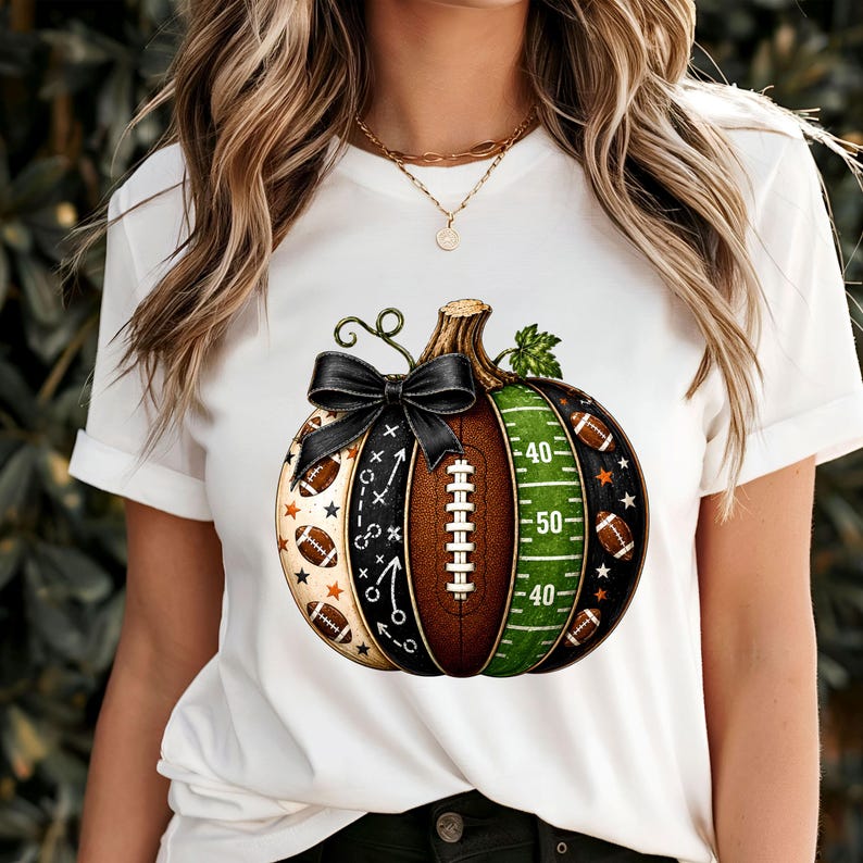 May include: A white t-shirt featuring a graphic of a pumpkin designed with football-themed elements. The pumpkin includes sections with football laces, a green field with yard lines, play diagrams, and small footballs, topped with a black bow.