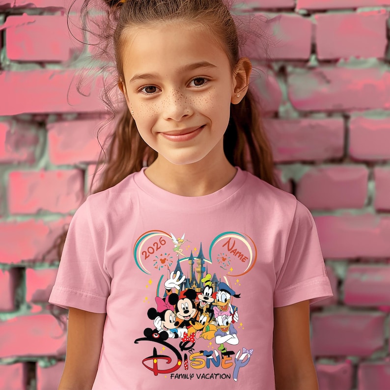 May include: Pink t-shirt featuring a Disney family vacation design. The graphic includes Mickey and Minnie Mouse, Donald and Daisy Duck, Goofy, and Pluto. The shirt has the text "2026" and "Name" in a Mickey Mouse ear design.