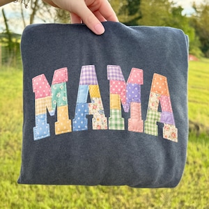 May include: A navy blue sweatshirt with the word "MAMA" appliqued across the front. The letters are made of colorful patchwork fabric in various patterns, including polka dots, floral, and gingham.