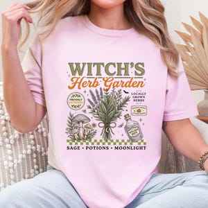 May include: A light pink t-shirt featuring a graphic print with the text "WITCH'S Herb Garden". The design includes illustrations of a bundle of herbs, mushrooms, a glass bottle labeled "WITCHES BREW", and text reading "FRESHLY GATHERED", "LOCALLY GROWN HERBS", "AUTUMN PICKED", "SAGE", "POTIONS", and "MOONLIGHT".