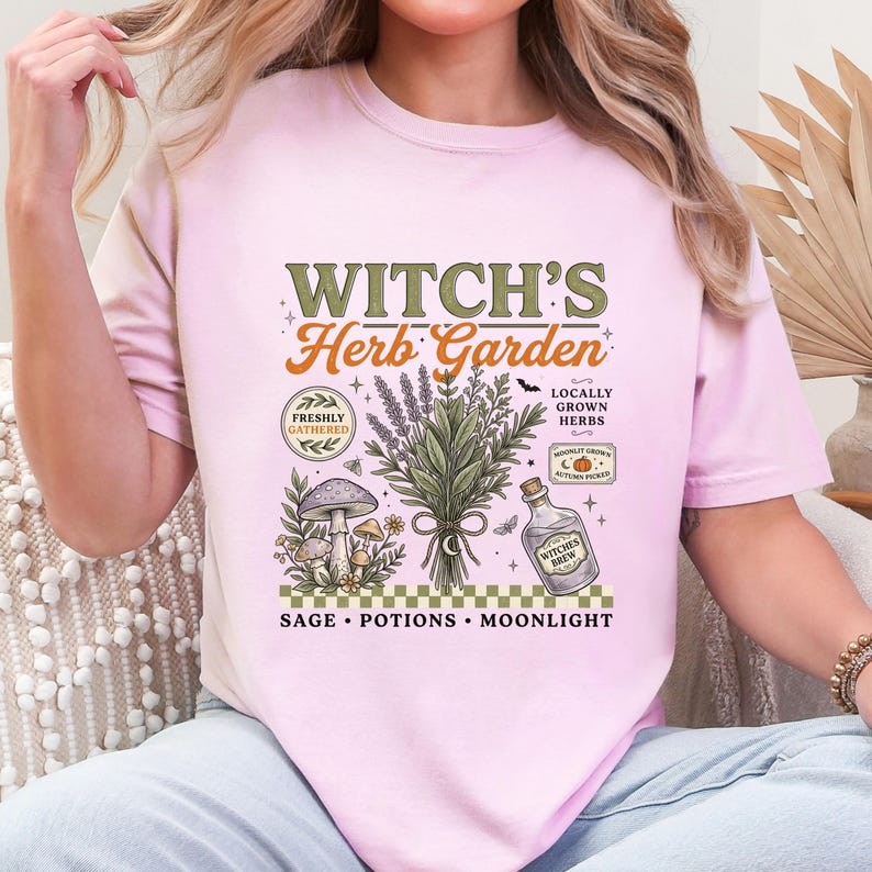 May include: A light pink t-shirt featuring a graphic print with the text "WITCH'S Herb Garden". The design includes illustrations of a bundle of herbs, mushrooms, a glass bottle labeled "WITCHES BREW", and text reading "FRESHLY GATHERED", "LOCALLY GROWN HERBS", "AUTUMN PICKED", "SAGE", "POTIONS", and "MOONLIGHT".