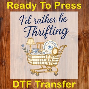 May include: A DTF transfer design featuring a shopping cart filled with vintage items like a lamp, teacup, and plates. The text "I'd rather be Thrifting" is in elegant script, with "Ready To Press" and "DTF Transfer" in yellow.