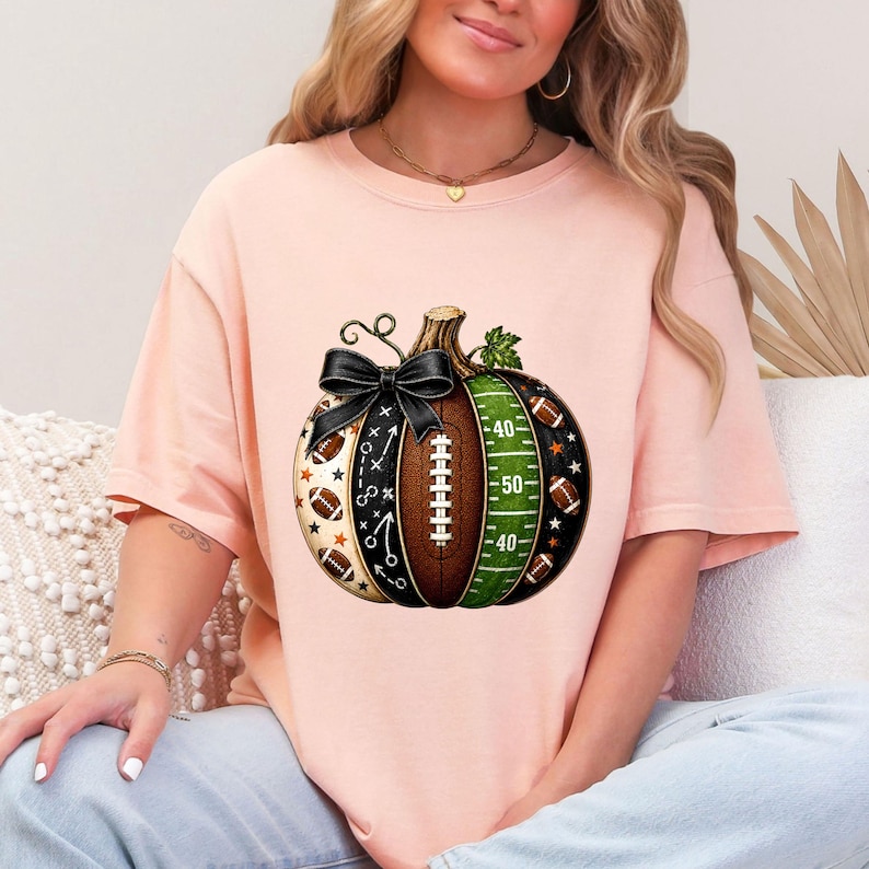 May include: A light peach-colored t-shirt featuring a central graphic of a pumpkin designed with American football elements. The pumpkin includes a black bow, a brown football texture, a green field with yard lines, and small football icons.