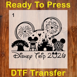 May include: Black DTF transfer with a Disney Trip 2026 design. The design includes Mickey Mouse ears with fireworks, a castle, a tree, and other Disney icons. The words "Ready To Press" and "DTF Transfer" are also included.