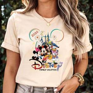 May include: A beige t-shirt with a vibrant Disney graphic. The design showcases Mickey and Minnie Mouse, Goofy, Donald Duck, and other characters. The word "Disney" is displayed with "Family Vacation" underneath, and the year "2026" is also visible.