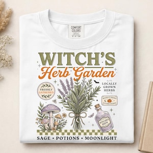 May include: A white t-shirt featuring a graphic print with the text "WITCH'S Herb Garden". The design includes illustrations of a bundle of herbs, a purple mushroom, a bottle labeled "WITCHES BREW", and a small pumpkin, all above a checkered border.