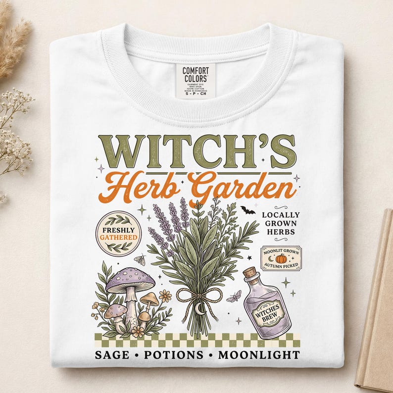 May include: A white t-shirt featuring a graphic print with the text "WITCH'S Herb Garden". The design includes illustrations of a bundle of herbs, a purple mushroom, a bottle labeled "WITCHES BREW", and a small pumpkin, all above a checkered border.