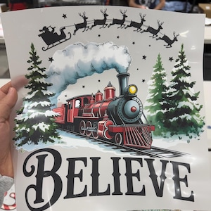Peut inclure: Graphique de No&euml;l avec un train &agrave; vapeur rouge, le tra&icirc;neau du P&egrave;re No&euml;l tir&eacute; par des rennes et des sapins. Le mot "BELIEVE" est &eacute;crit en gras, cr&eacute;ant une ambiance festive.