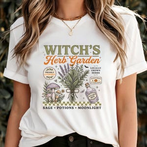 May include: A white t-shirt featuring a graphic print with the text "WITCH'S Herb Garden" and "SAGE &bull; POTIONS &bull; MOONLIGHT". The design includes illustrations of a bundle of herbs, mushrooms, a bottle labeled "WITCHES BREW", and small decorative elements.