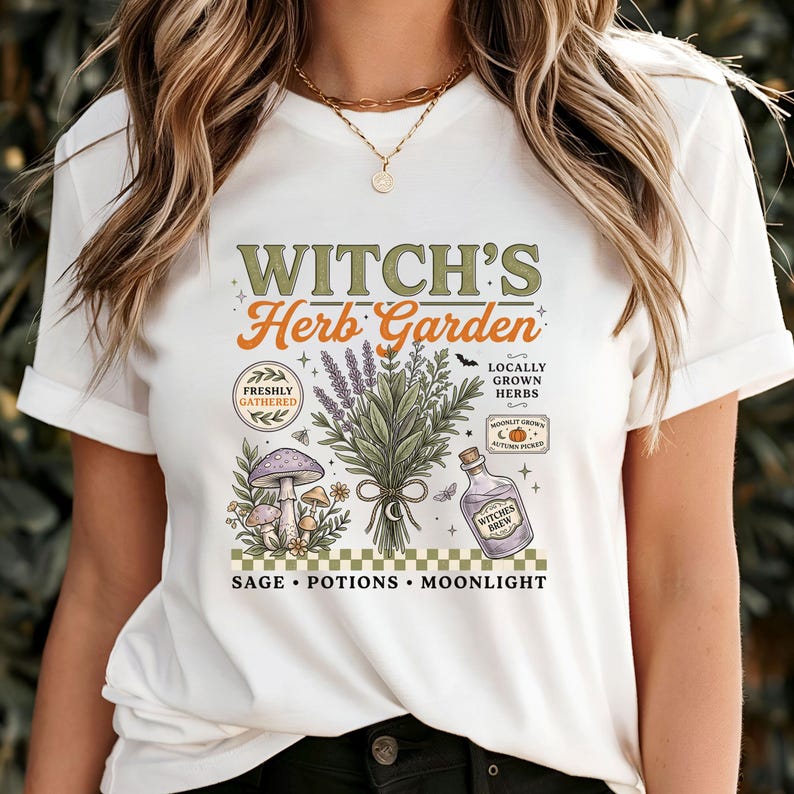 May include: A white t-shirt featuring a graphic print with the text "WITCH'S Herb Garden" and "SAGE &bull; POTIONS &bull; MOONLIGHT". The design includes illustrations of a bundle of herbs, mushrooms, a bottle labeled "WITCHES BREW", and small decorative elements.
