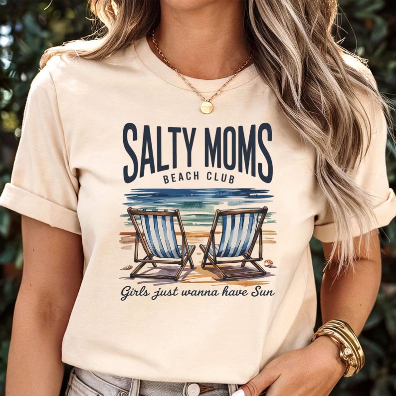 May include: A cream-coloured t-shirt featuring the text "SALTY MOMS BEACH CLUB" above a beach scene with two deckchairs. The phrase "Girls just wanna have Sun" is printed below. The shirt has short sleeves and a relaxed fit.