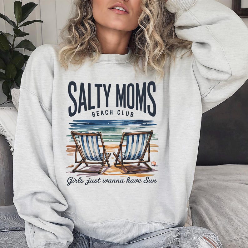 May include: A pale grey sweatshirt featuring the text "SALTY MOMS BEACH CLUB" above a beach scene with two blue and white striped deckchairs. The phrase "Girls just wanna have Sun" is printed below the beach scene.