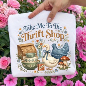 May include: White graphic t-shirt with the text "Take Me To The Thrift Shop." The design includes a bread box, bowls, a duck with a blue bow, a blue hen, mushrooms, and flowers. The shirt is held up by a hand.