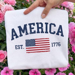 May include: A white t-shirt featuring the dark blue distressed text AMERICA arched over a graphic of the United States flag. To the left of the flag is the text EST. and to the right is the number 1776, both in a dark blue distressed font.