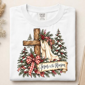 Puede incluir: Una camiseta blanca con un gráfico de una cruz de madera cubierta con tela blanca, rodeada de pinos nevados, bayas rojas y lazos a cuadros rojos y blancos. Una cinta en la parte inferior muestra el texto Jesus is the Reason.