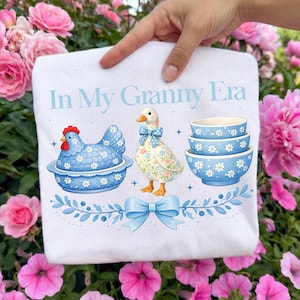 May include: White sweatshirt with the text "In My Granny Era" in light blue. The design features a blue chicken in a bowl, a floral duck with a bow, and three blue bowls with daisy patterns. A blue bow and floral elements complete the design.
