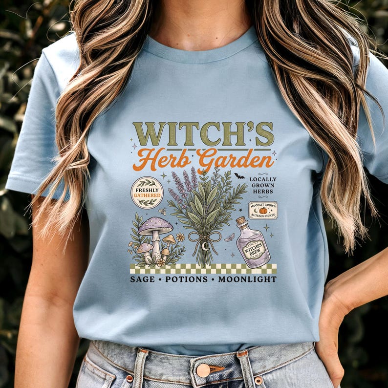 May include: A light blue t-shirt featuring a graphic print with the text Witch's Herb Garden. The design includes a bouquet of herbs, mushrooms, a potion bottle, and small icons like a pumpkin and moon. The bottom text reads Sage, Potions, Moonlight.