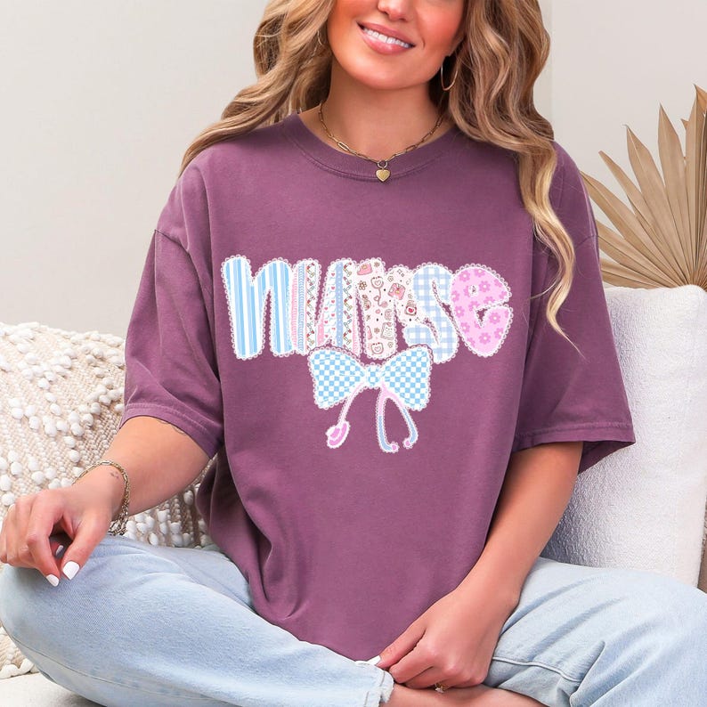May include: A mauve-colored t-shirt with the word "NURSE" in a patchwork design. The letters are decorated with blue and white stripes, pink floral patterns, and a blue gingham bow with a stethoscope.