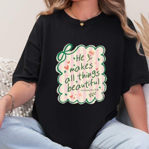 May include: Black t-shirt featuring a colorful graphic with the text "He makes all things beautiful" in a whimsical font. The design includes hearts, flowers, and stars. The shirt is a casual, everyday wear item.
