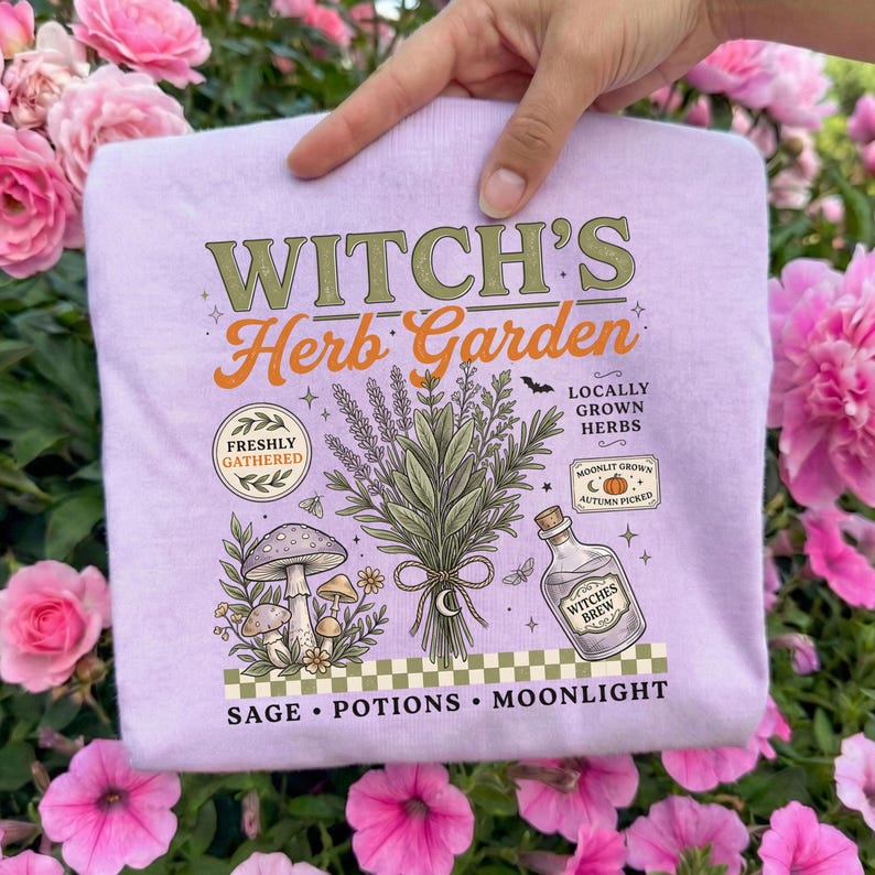 May include: A light purple t-shirt featuring a graphic with the text Witch's Herb Garden, Freshly Gathered, Locally Grown Herbs, Moonlit Grown Autumn Picked, and Sage, Potions, Moonlight. The design includes a bouquet of herbs, mushrooms, and a bottle.